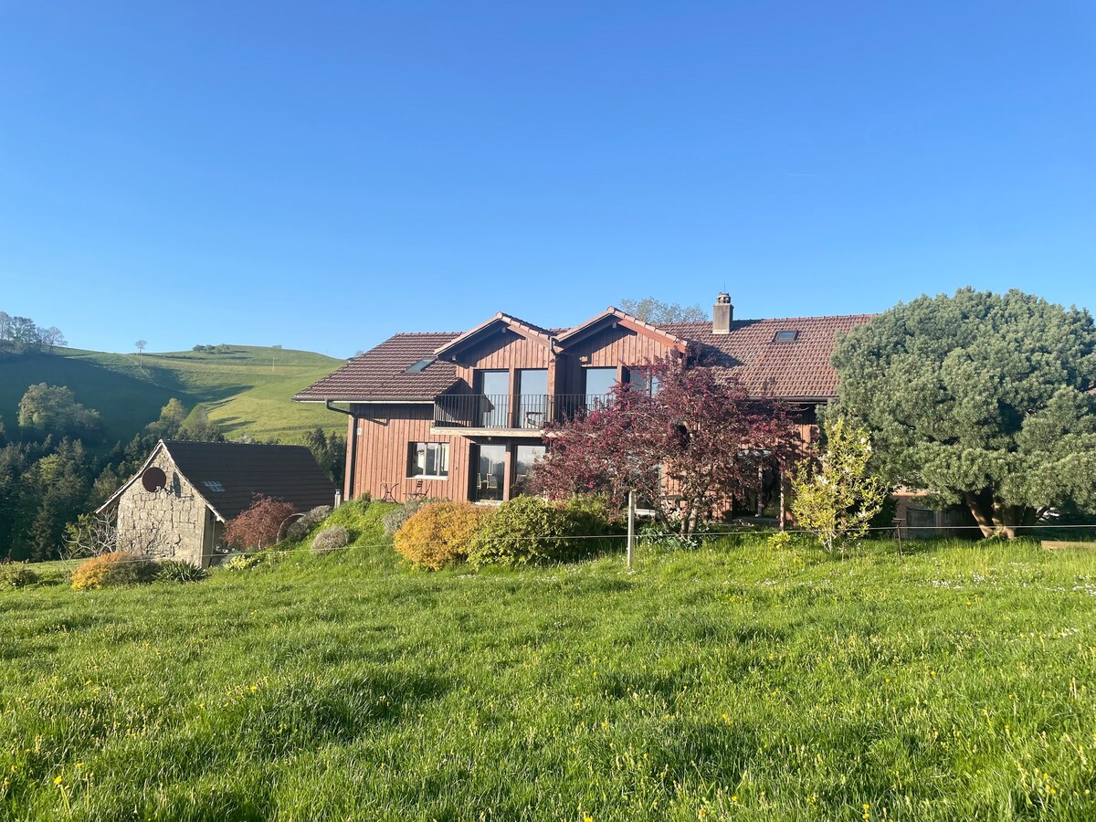 A two-story wooden house is set against a blue sky, with green hills in the background. Surrounding the house, a variety of trees and flowering plants add color to the lush grassy area, enhancing the connection to the natural landscape.