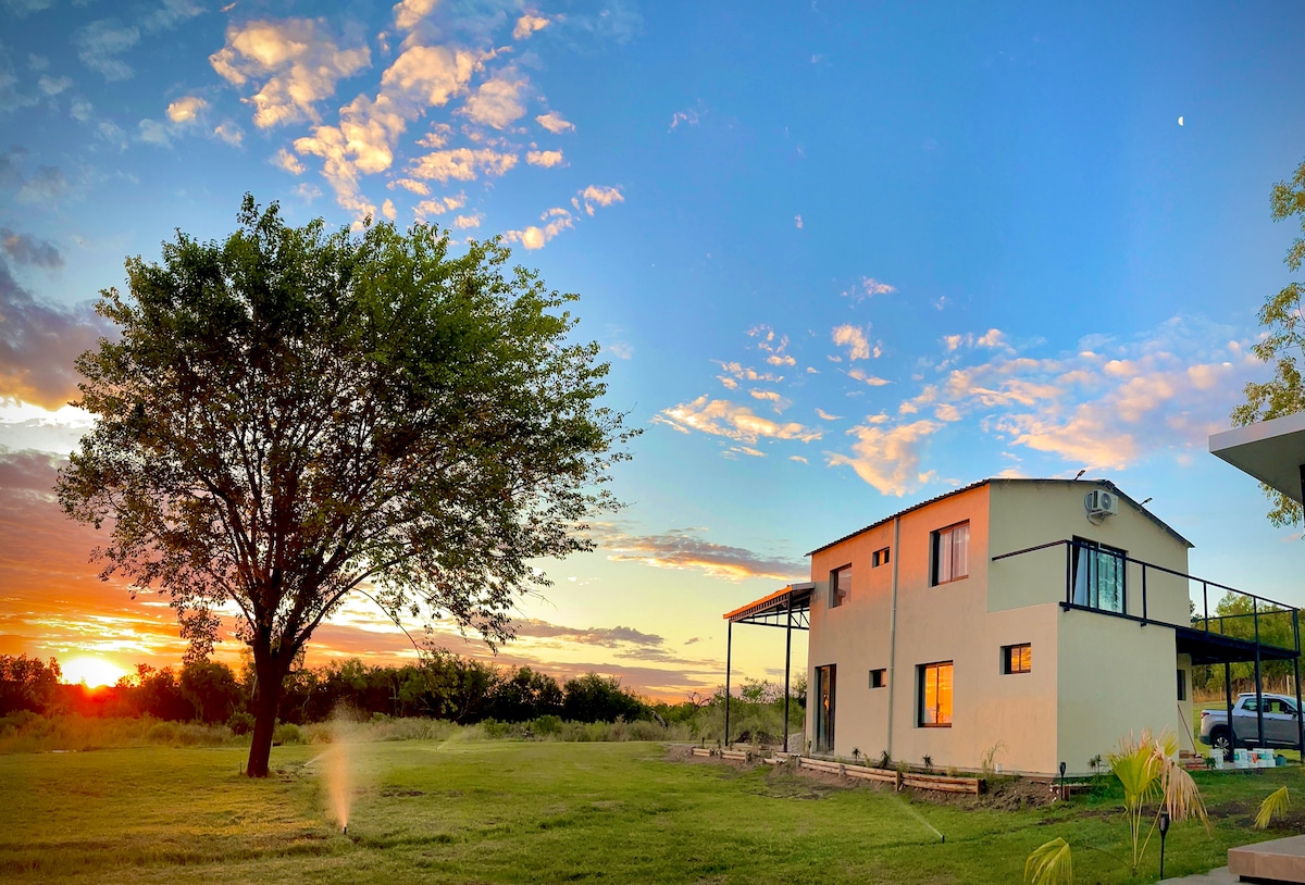 A serene landscape features a two-story house with large windows and a covered porch. A sprawling green lawn surrounds the property, complemented by a substantial tree. The sunset casts warm hues across the sky, creating a peaceful atmosphere.