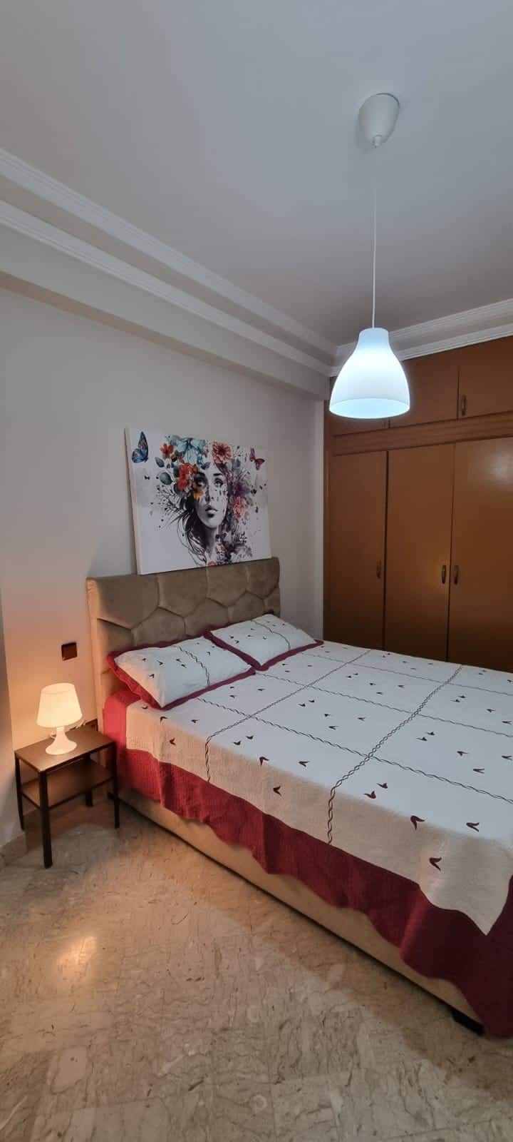 A cozy bedroom is shown, featuring a spacious bed dressed in patterned linens. A soft bedside lamp provides gentle lighting next to a wall adorned with floral artwork. Ample storage is available in the wooden wardrobe located on the opposite side.