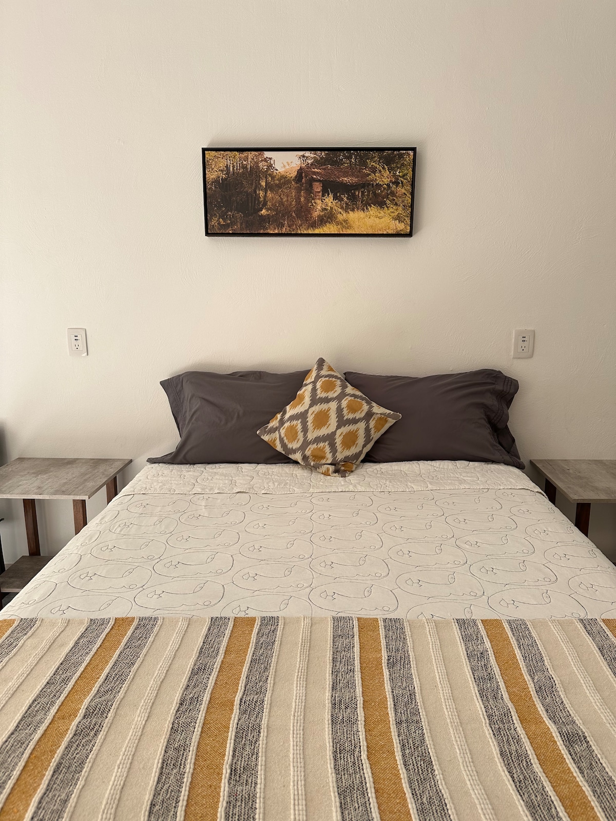 A spacious bed is featured with neutral-toned bedding and a decorative pillow. Two wooden nightstands are positioned on either side. A wall-mounted photograph of an outdoor scene is displayed above the bed, adding a touch of nature to the room.