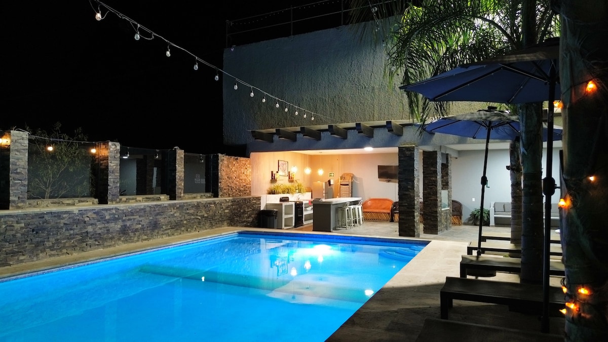 A tranquil outdoor space features a lit swimming pool surrounded by a stone terrace. A covered seating area is visible, accompanied by an outdoor kitchen. Strings of lights create a soft illumination, enhancing the serene ambiance of the nighttime setting.