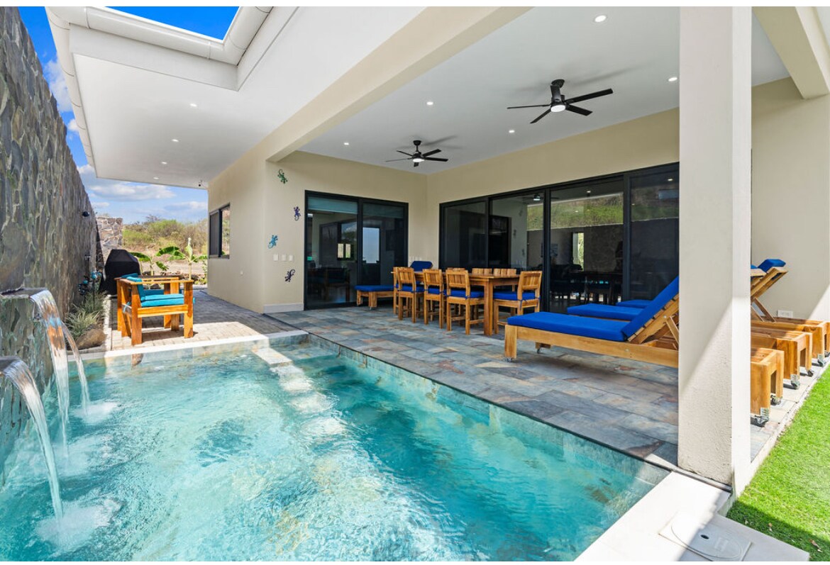 A large private patio features a refreshing pool with cascading water. Comfortable loungers and a spacious dining table invite relaxation and outdoor meals. Ceiling fans provide a cooling breeze, while large glass doors connect to the interior, enhancing the open and airy feel of the space.