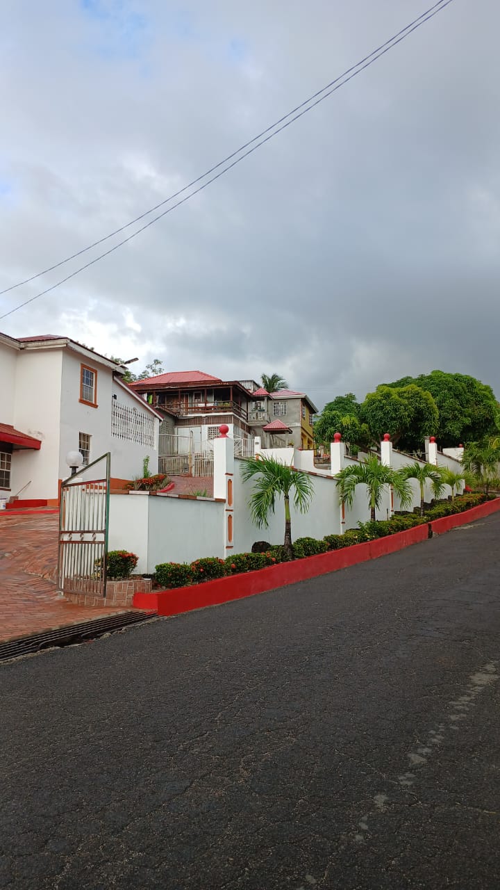 A well-maintained exterior view of a property set along a gently sloping road. The fence lined with vibrant greenery and decorative posts leads to a multi-story building with red roofing. A welcoming pathway can be seen, inviting guests to explore.