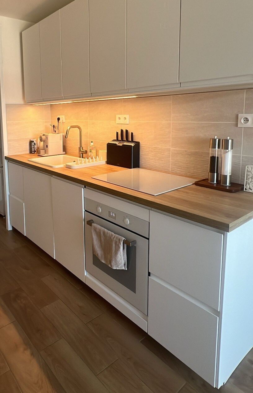 A modern kitchen features a minimalist design with white cabinetry and a wood countertop. An oven, sink, and cooktop are visible, accompanied by a range of kitchen utensils. Under-cabinet lighting illuminates the space, enhancing its functionality and clean aesthetic.