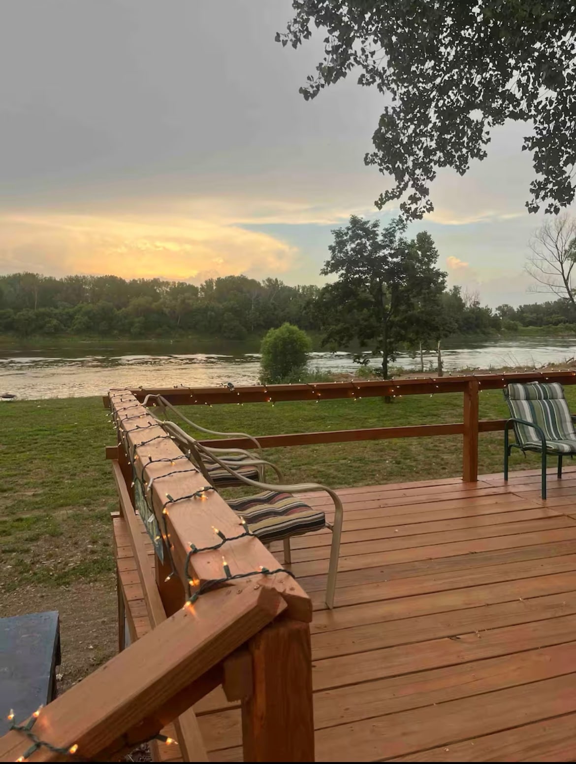 A wooden deck featuring two chairs, adorned with soft lighting, overlooks a serene river and lush greenery. The setting sun casts a warm glow across the landscape, enhancing the natural beauty of the riverfront location.