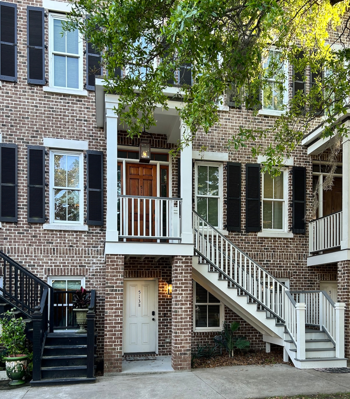 The exterior of a brick townhouse is showcased, featuring a welcoming front entrance framed by tall windows and black shutters. A set of stairs leads up to a covered porch with a wooden front door, surrounded by lush greenery and outdoor steps.