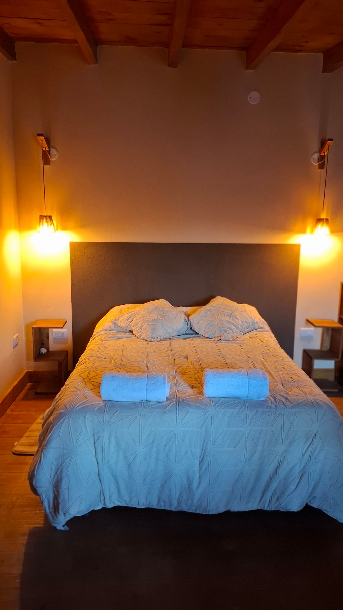 A spacious bed is situated against a textured wall, complemented by warm bedside lighting. Two white towels are neatly placed on the bed, accentuating the cozy atmosphere of the room. The wooden nightstands on either side provide a natural touch.