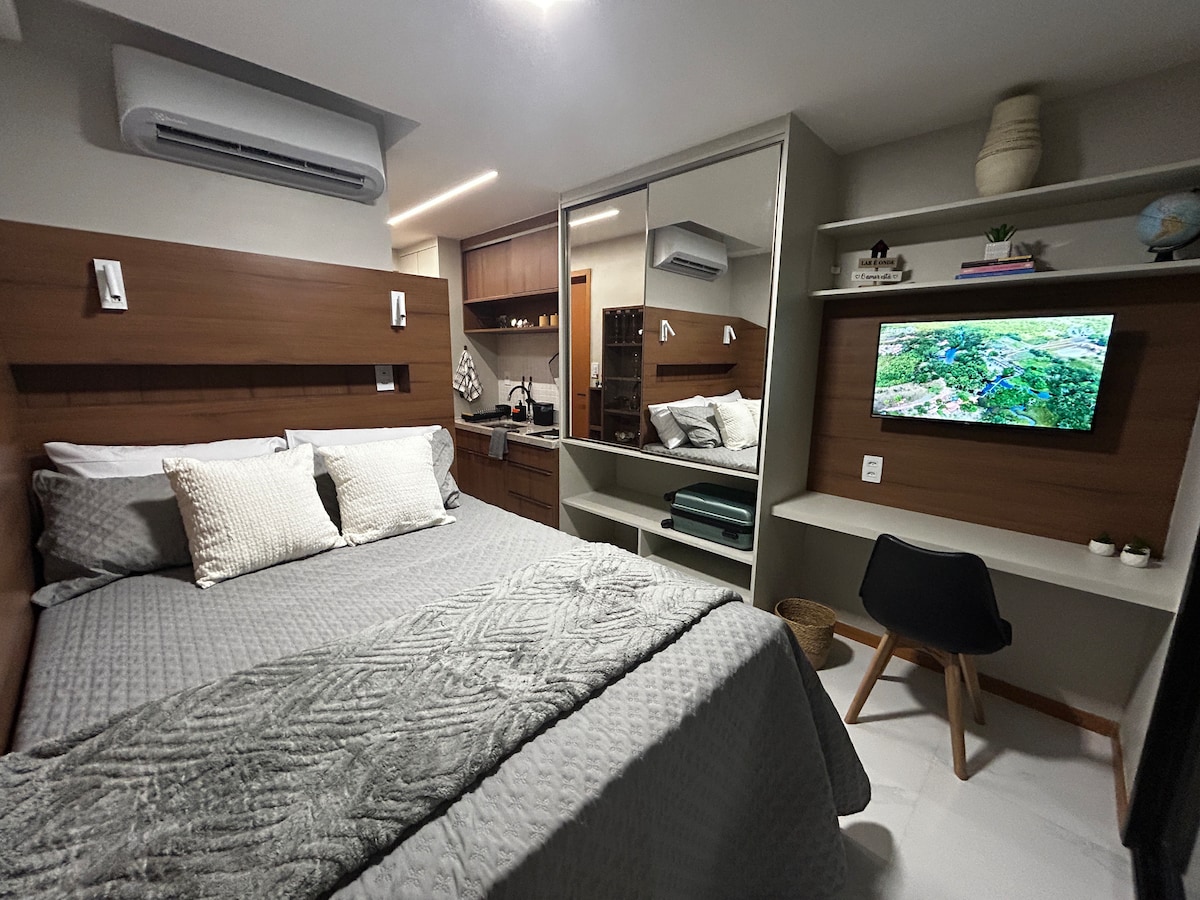 A private sleeping area features a spacious bed with light grey bedding and multiple decorative pillows. A mounted television is positioned across from the bed, with a small desk and chair nearby. Soft lighting and a mirrored wardrobe enhance the bright, modern atmosphere.