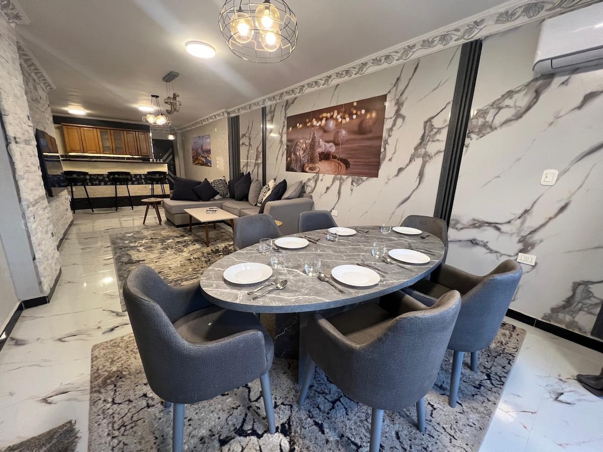 A spacious dining area features a large oval table set for six, adorned with plates and glassware. Stylish grey upholstered chairs surround the table. The room is enhanced by elegant marble walls and modern artwork, providing a modern yet comfortable dining experience.