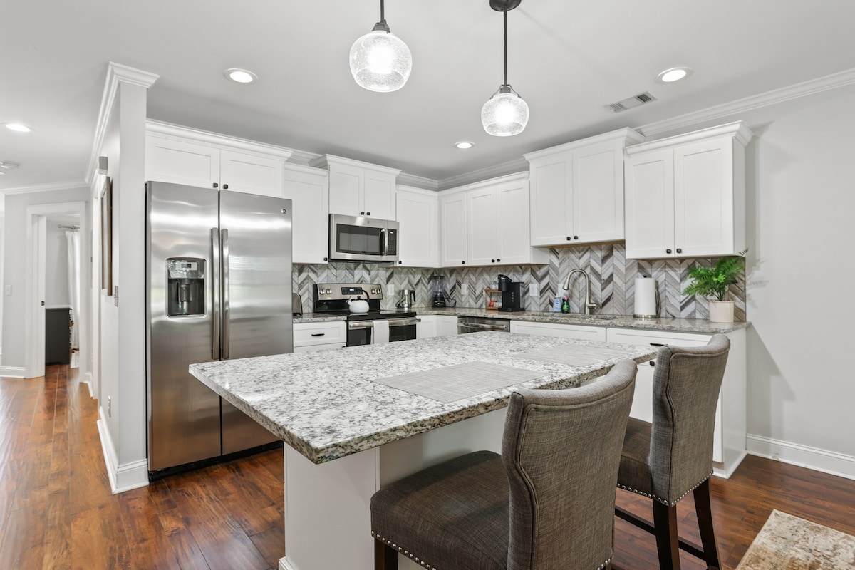 A modern kitchen is showcased, featuring stainless steel appliances and granite countertops. A large island with seating for two is present, complemented by stylish pendant lighting. Decorative tile backsplash enhances the aesthetic, while light-colored cabinetry provides ample storage.