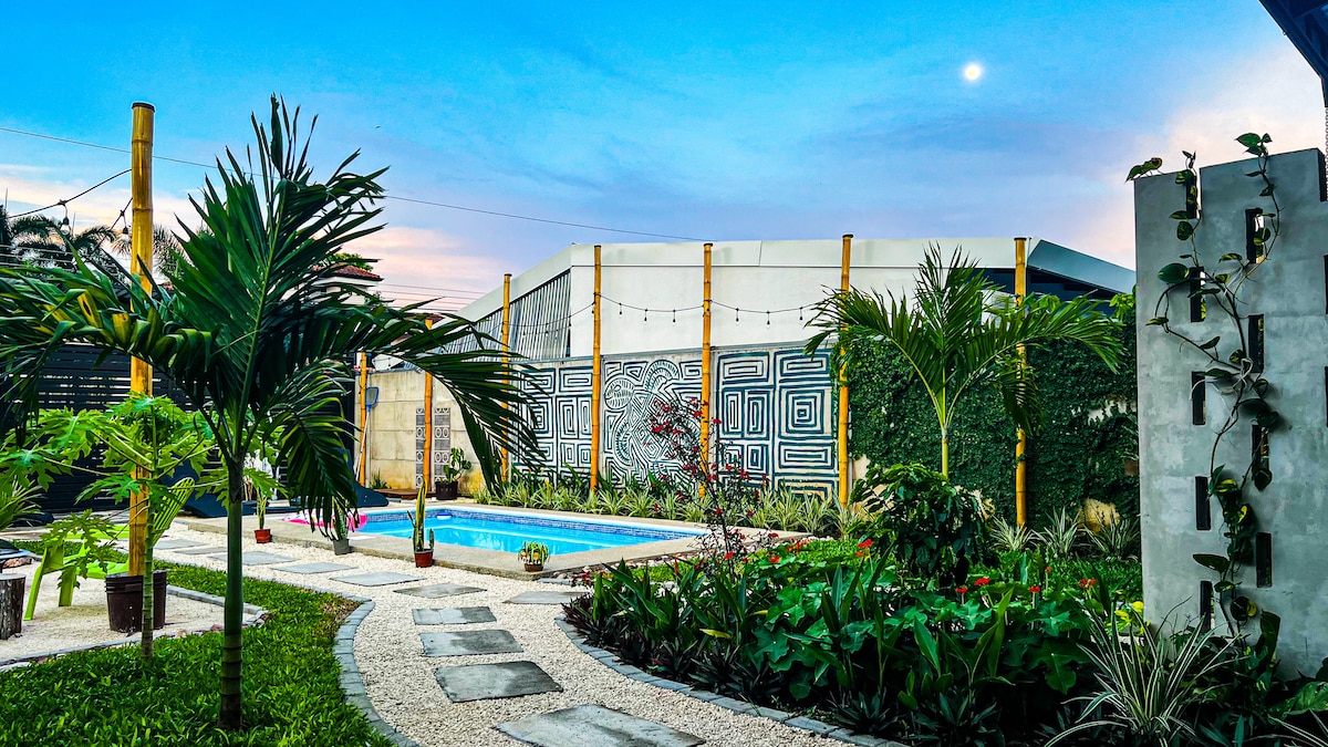 A lush garden is displayed, featuring tropical plants and a well-maintained pathway leading to a sparkling swimming pool. Decorative walls provide visual interest, while an evening sky hints at a tranquil atmosphere. Lights are softly illuminated, enhancing the serene outdoor space.