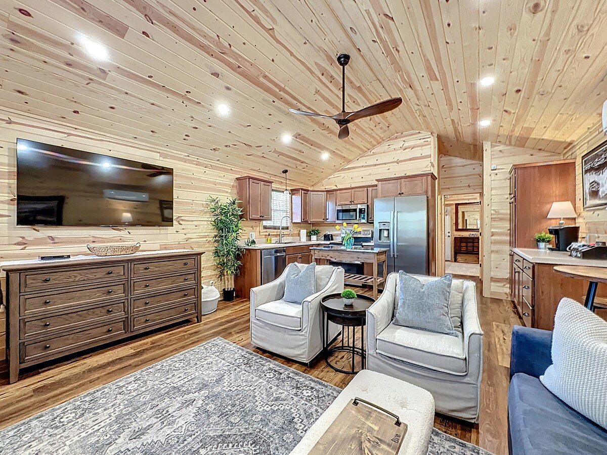 A spacious living area features a large flat-screen TV mounted on a wooden wall. Comfortable seating is arranged around a coffee table, accompanied by a textured area rug. The open layout includes a view of a modern kitchen with wooden cabinetry and stainless steel appliances.