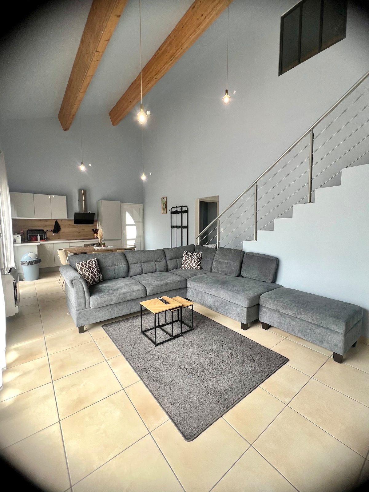 A spacious living area is featured with high ceilings and wooden beams. A gray sectional sofa is positioned on a light tiled floor, accompanied by a small black coffee table. Soft light from hanging fixtures illuminates the room, while a staircase leads to an upper level.
