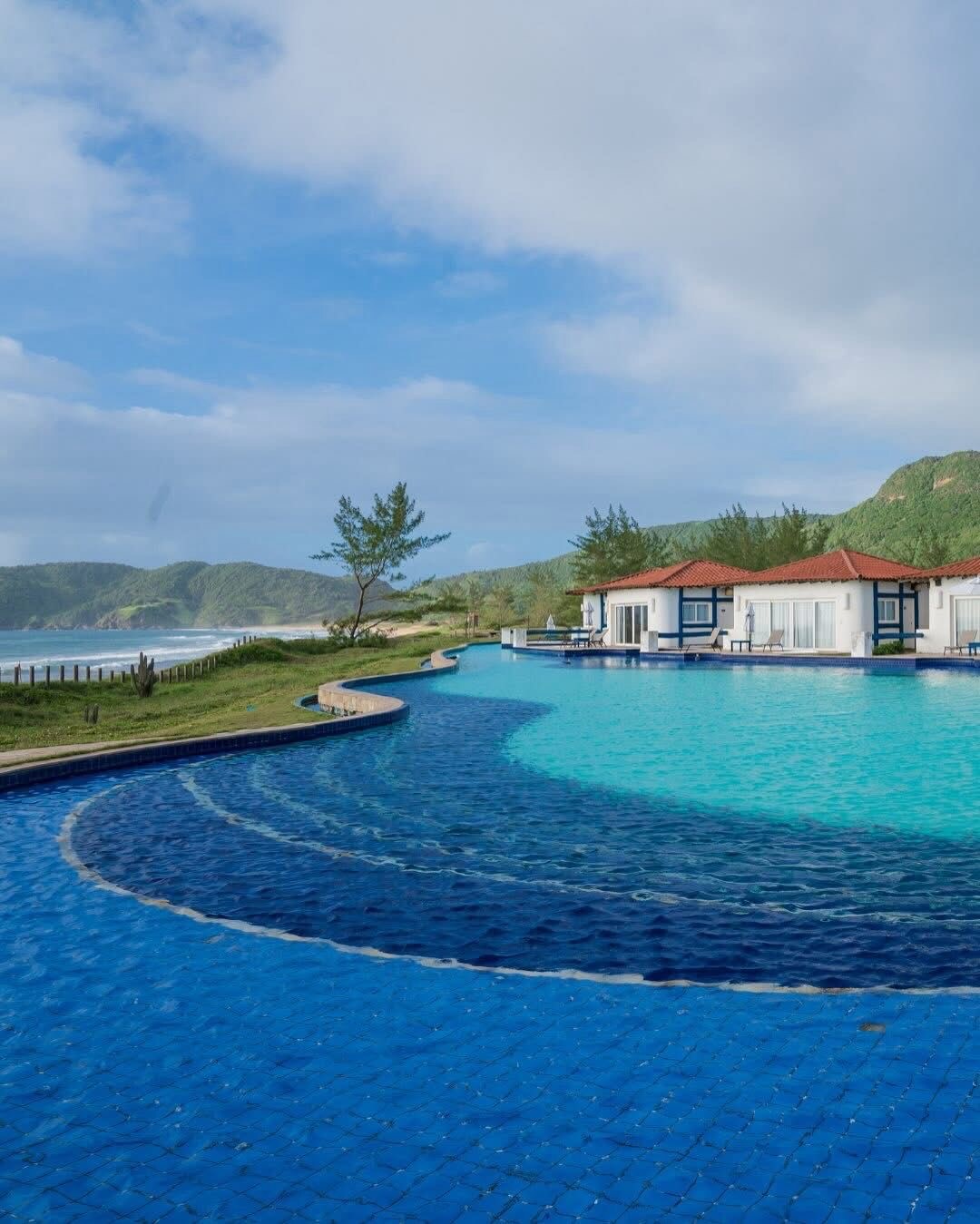 An expansive swimming pool glistens with rich blue tiles, curving gracefully along the landscape. Surrounding the pool are charming white cottages with red roofs, complemented by lush greenery and rolling hills in the background under a clear sky.