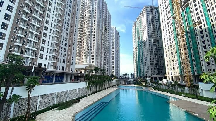 A large swimming pool is visible, surrounded by modern high-rise apartment buildings. The pool is framed by landscaped gardens, featuring palm trees and manicured greenery. The clear blue sky reflects on the water's surface, creating a serene atmosphere.