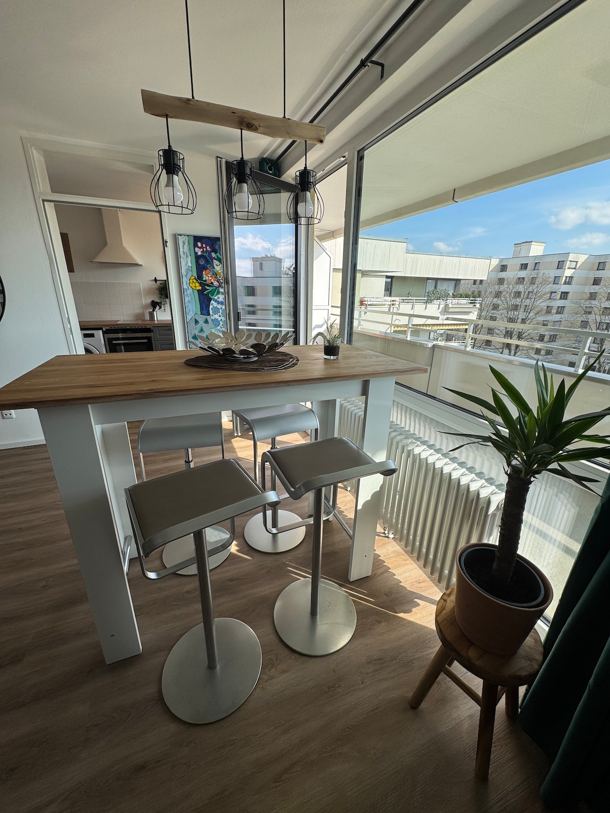 A dining area is highlighted by a wooden table with three bar stools. Large windows provide natural light and showcase an outdoor view. A potted plant adds a touch of greenery in the corner, while unique light fixtures hang above the table.