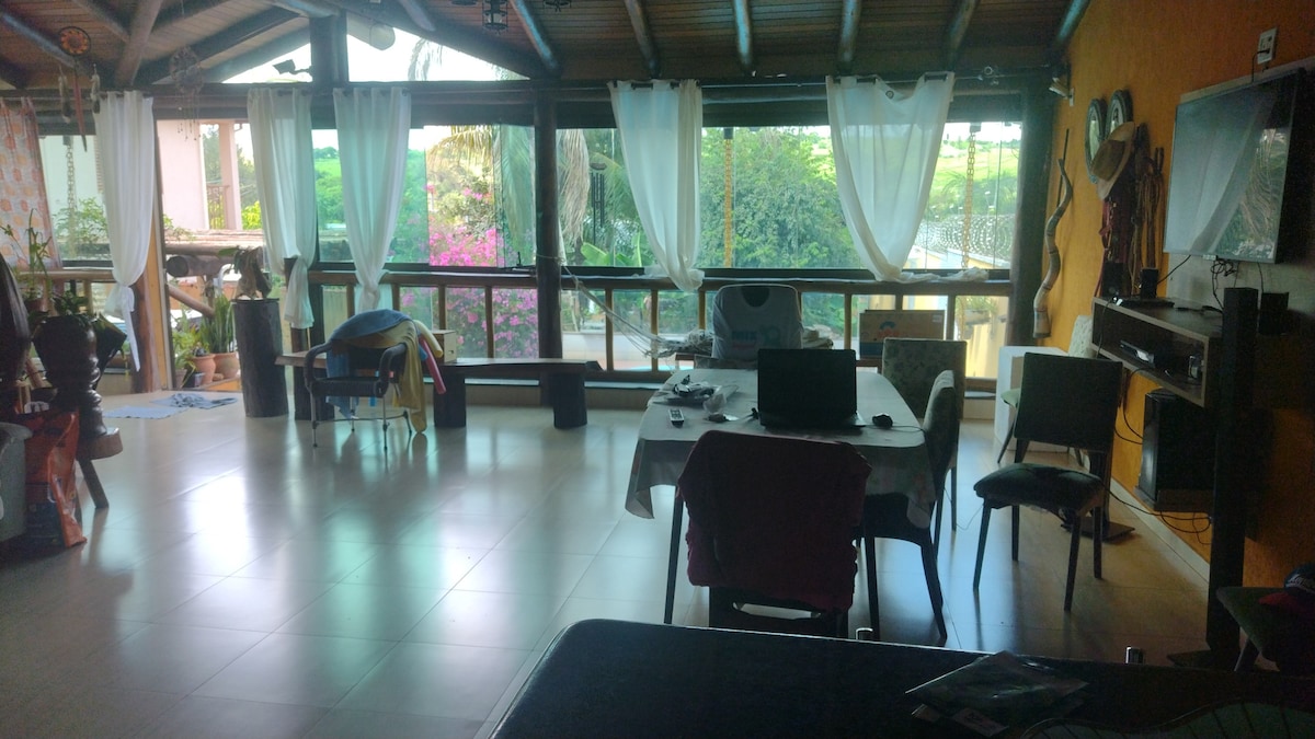 An open living area is seen, featuring large glass windows that provide views of the garden. A dining table is positioned alongside several chairs. Decorative plants and vibrant flowers are visible outside, contributing to a bright and airy atmosphere.