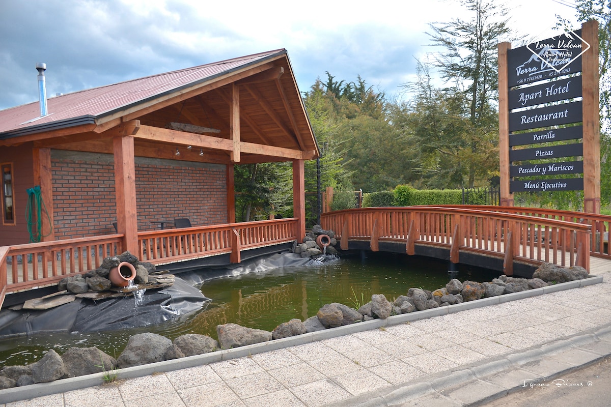 A rustic wooden structure features a welcoming entrance with a landscaped pond adorned with rocks and water features. A sign indicates the presence of an apart hotel and restaurant services. The surrounding area is dotted with greenery, offering a serene ambiance.