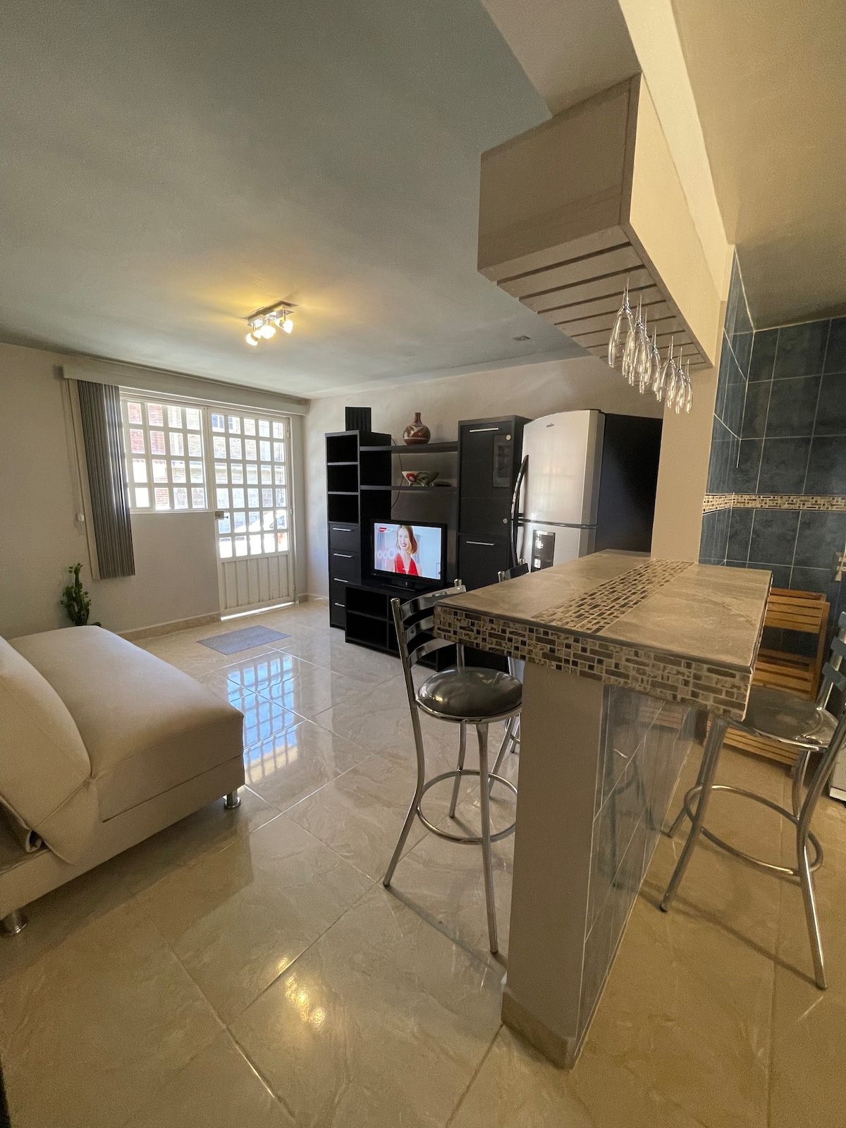 A comfortable living space features a light-colored sofa facing a media unit with a television. Natural light filters in through a grid-patterned window. Bar stools are arranged around a sleek counter, complemented by dark cabinetry and a silver refrigerator in the kitchen area.