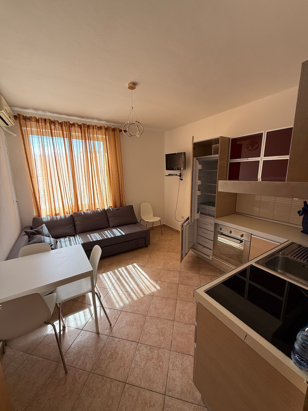 A cozy living area features a spacious gray sofa, a compact dining table with white chairs, and a small wall-mounted television. Light filters through sheer orange curtains, illuminating the tiled floor and highlighting the modern kitchen cabinetry and appliances.