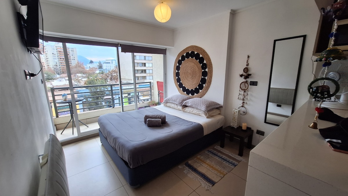 A spacious bedroom features a large bed with neutral bedding and a towel neatly placed on top. A decorative wall hanging adds visual interest. Natural light enters through the sliding glass door, which opens to a balcony overlooking the city. A minimalist bedside table completes the space.