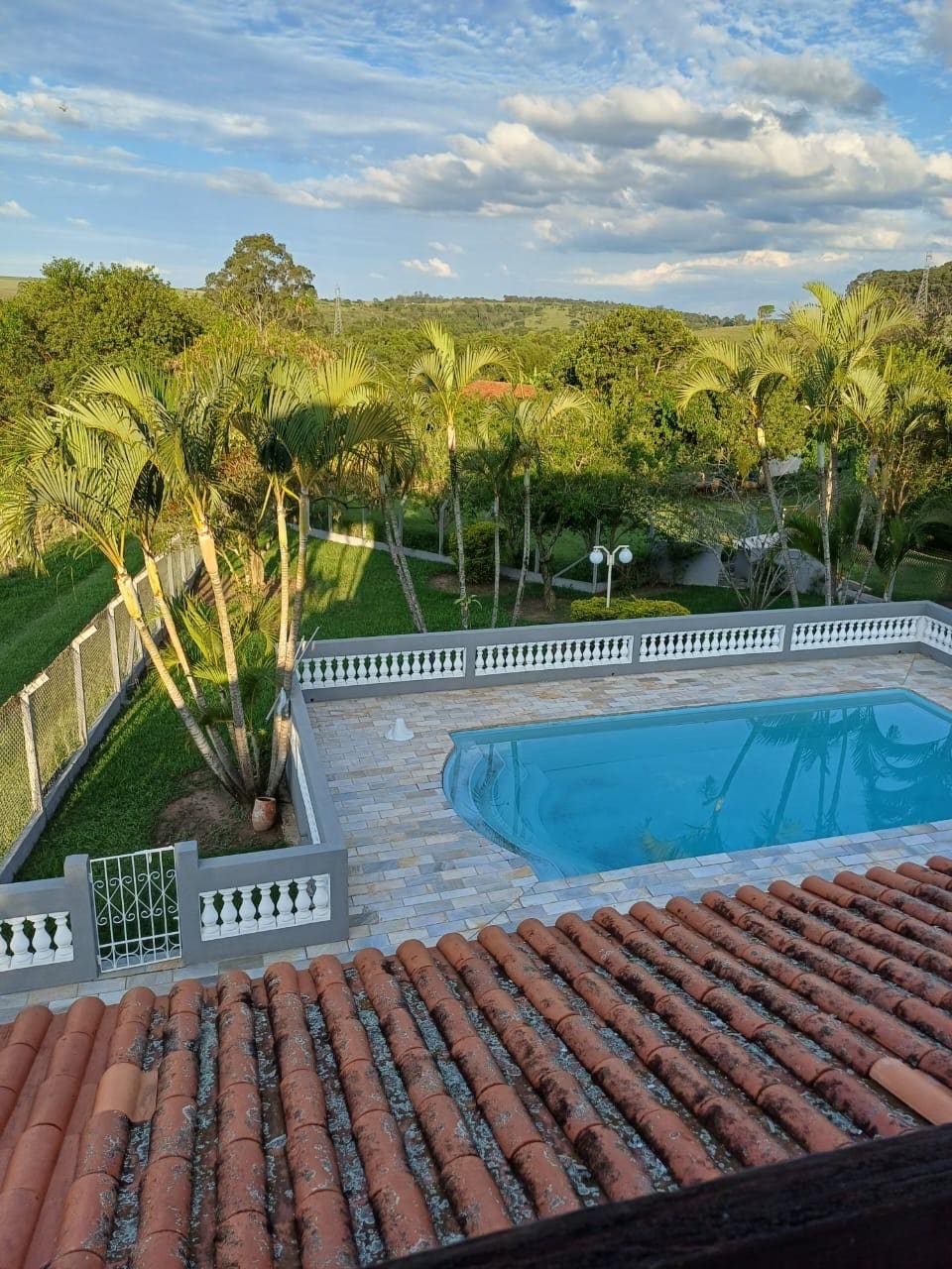 An aerial view of a tranquil swimming pool surrounded by lush greenery and palm trees. The pool features a clear blue water surface, and the area is paved with light-colored stone. Fencing and lighting elements are also visible in the serene outdoor environment.