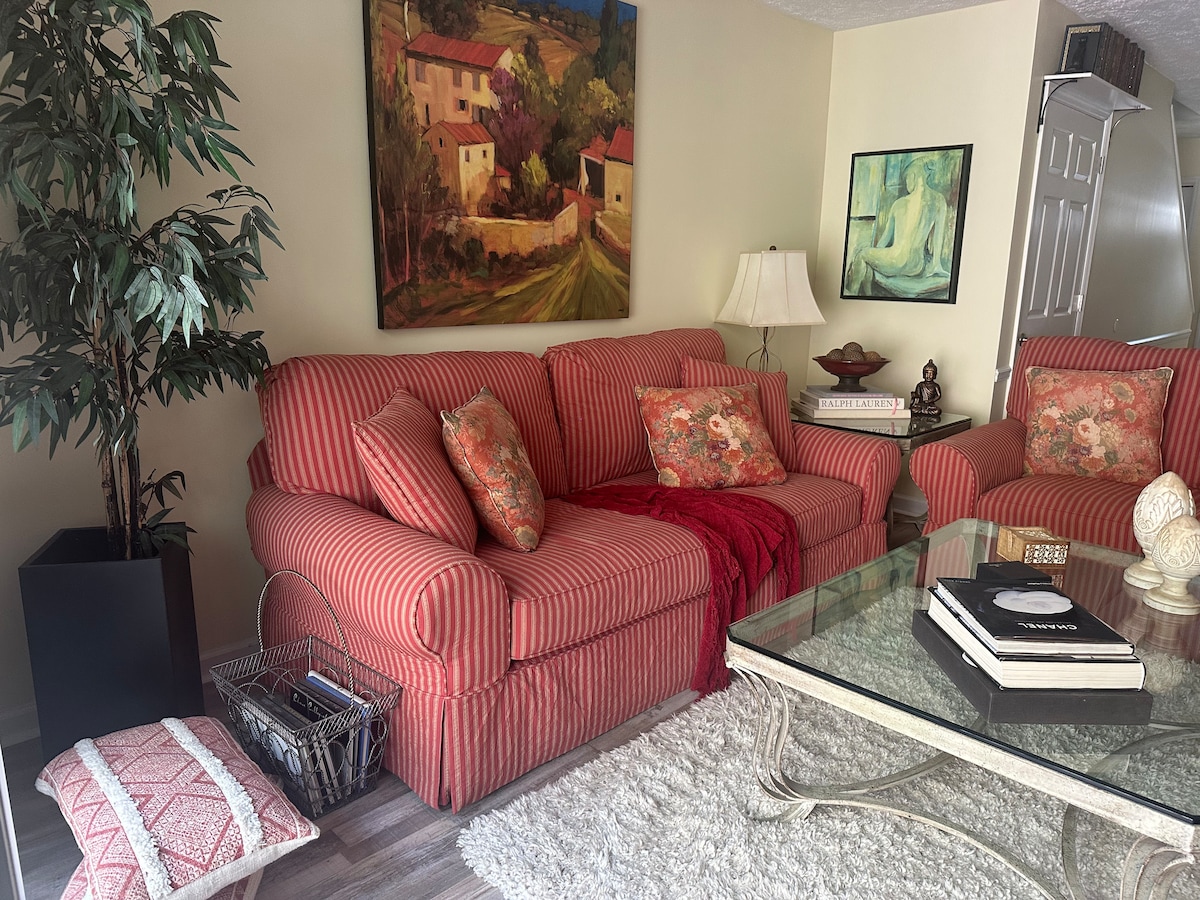 A comfortable living area is presented with a striped sofa adorned with decorative pillows. A glass coffee table is centered on a light-colored rug, with a small accent table featuring a lamp. A potted plant and artwork decorate the space, providing a relaxed ambiance.
