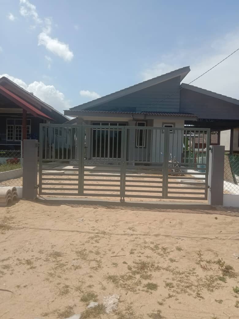 A grey gate leads to a modest house with a slanted roof and a covered porch area. Surrounding sandy terrain offers a natural, open feel. The entrance is bordered by a low fence, creating a welcoming boundary for the property.