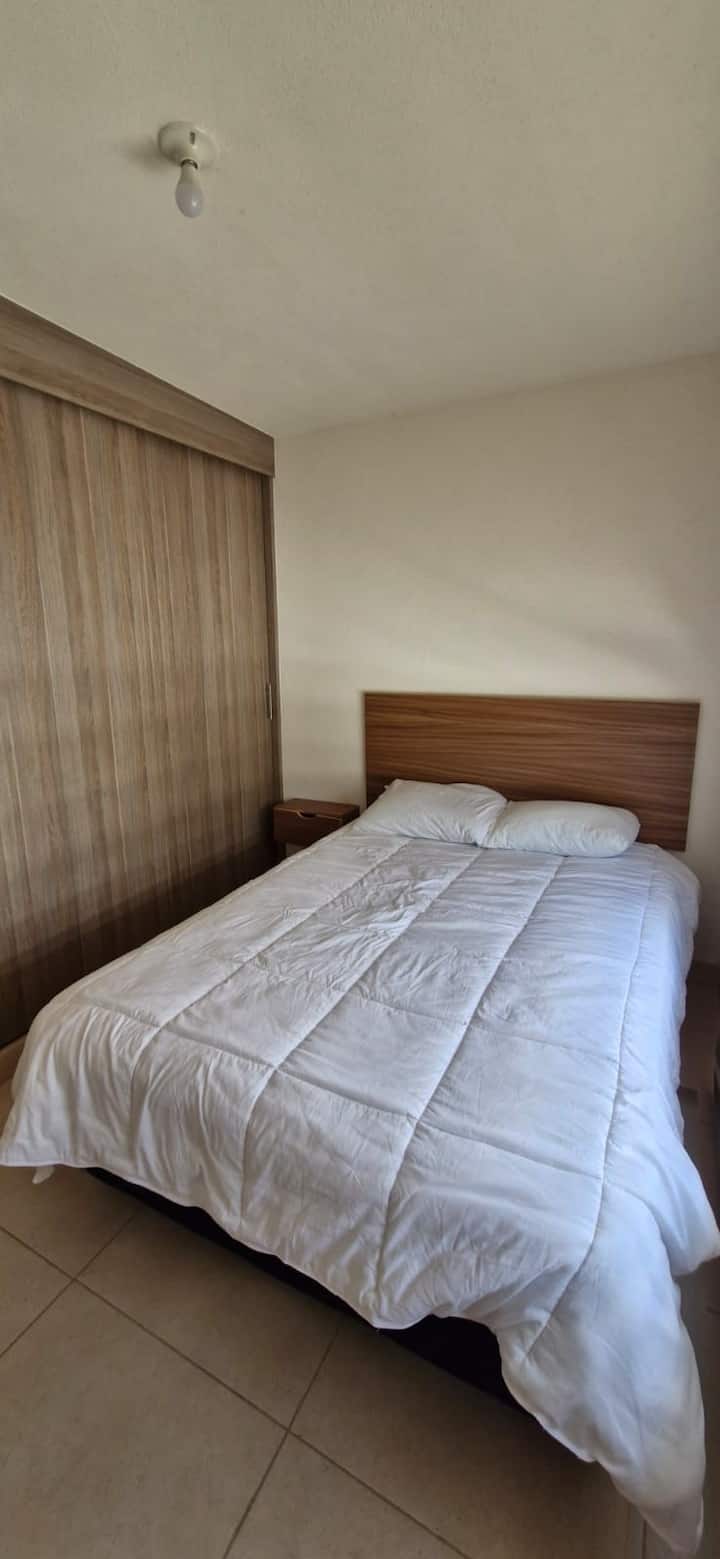 Cómoda recámara equipada con cama matrimonial, ropa de cama blanca y cabecera de madera. Destaca su amplio clóset de piso a techo con puertas corredizas, ideal para guardar todo tu equipaje y mantener el orden durante tu estancia.