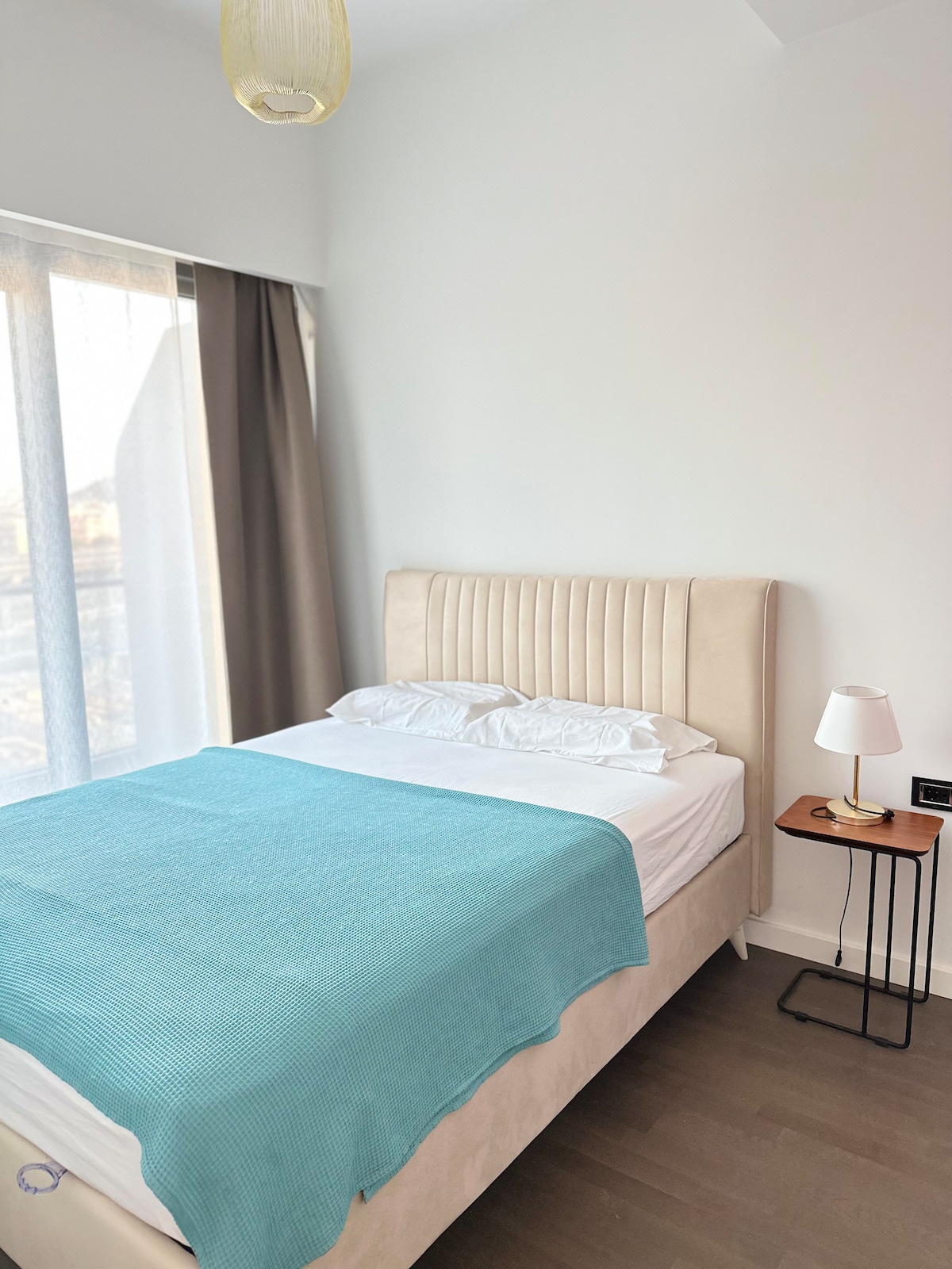 A comfortable sleeping area is arranged with a light-colored headboard and a spacious bed covered with a soft blue blanket. Natural light filters through sheer curtains, creating a serene ambiance. A small bedside table holds a minimalist lamp, providing additional lighting.