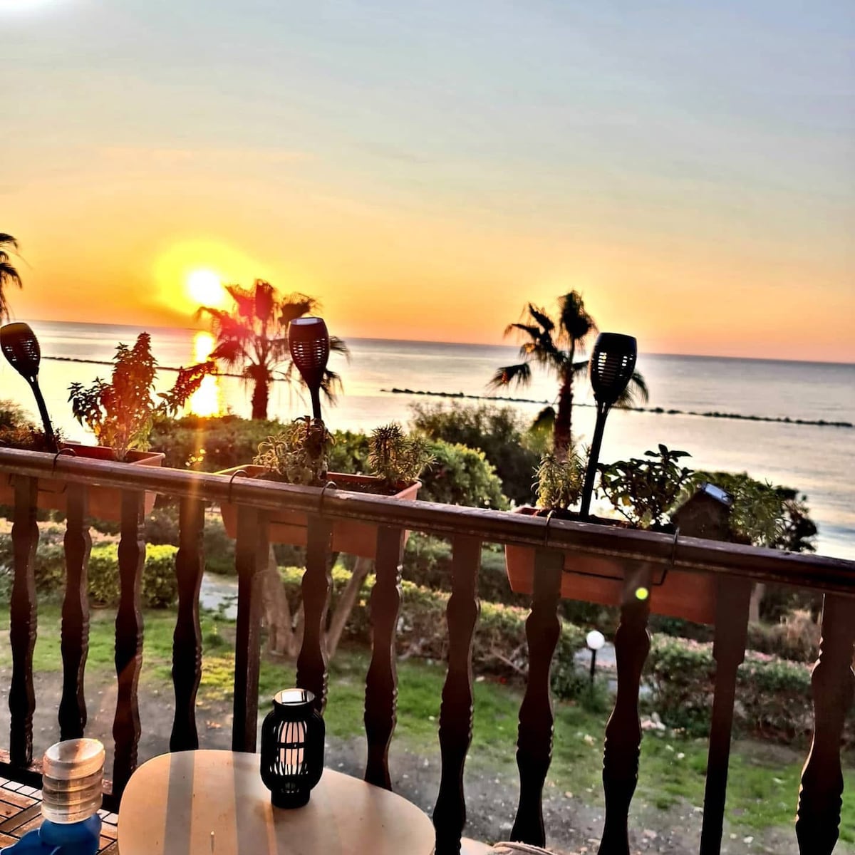 A seaside balcony is featured, showcasing potted plants and lanterns, with a vibrant sunset casting warm hues over the horizon. The calm sea and silhouetted palm trees are visible, providing a peaceful setting for morning coffee or evening relaxation.