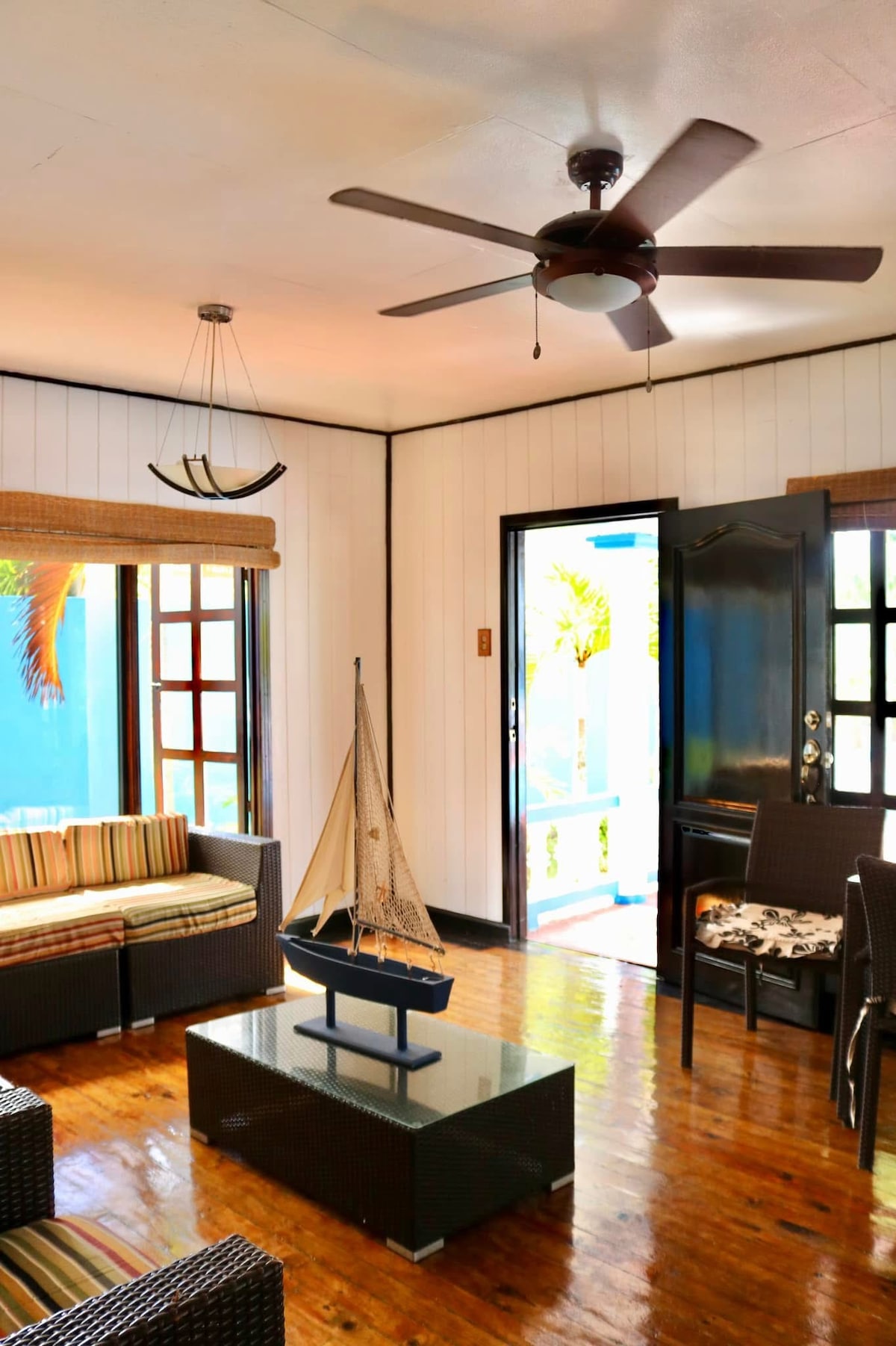 A bright living area features a ceiling fan and a wooden floor that reflects natural light. Comfortable seating is arranged around a rectangular coffee table, which holds a decorative sailboat. A doorway opens to an outdoor space, enhanced by views of greenery.