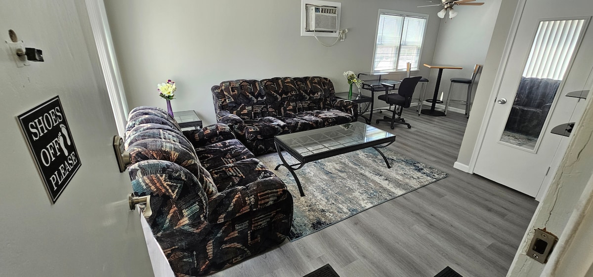 A living area is furnished with a pair of patterned sofas and a glass coffee table atop a soft rug. A small dining area with high chairs is visible in the background, complemented by a window allowing natural light. An air conditioning unit is mounted on the wall.