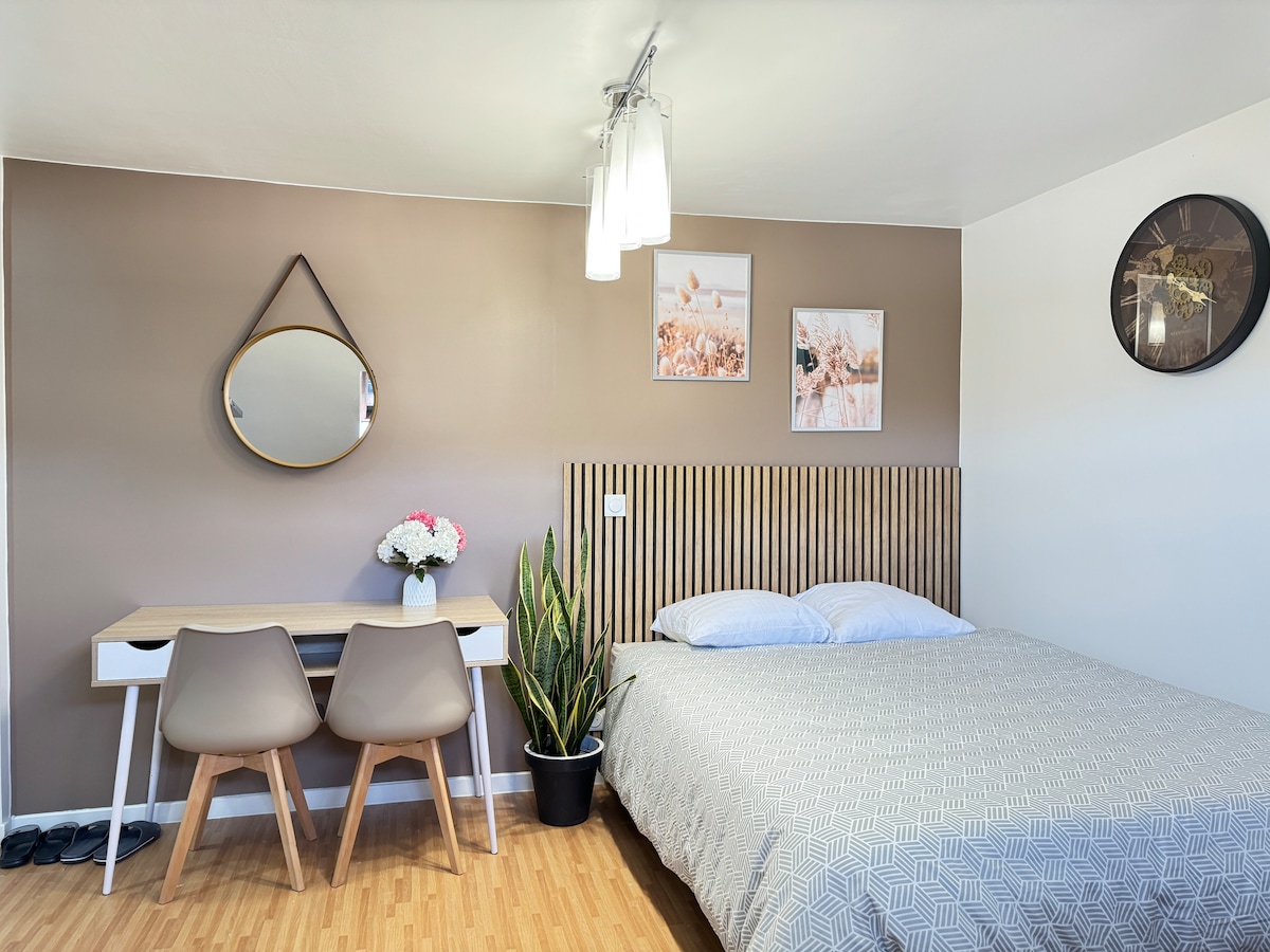 A room features a spacious bed with a textured grey coverlet. A wooden headboard adds warmth, while a round mirror hangs above a minimalist desk with two chairs. A plant in a pot brightens the corner, and framed art enhances the decor.