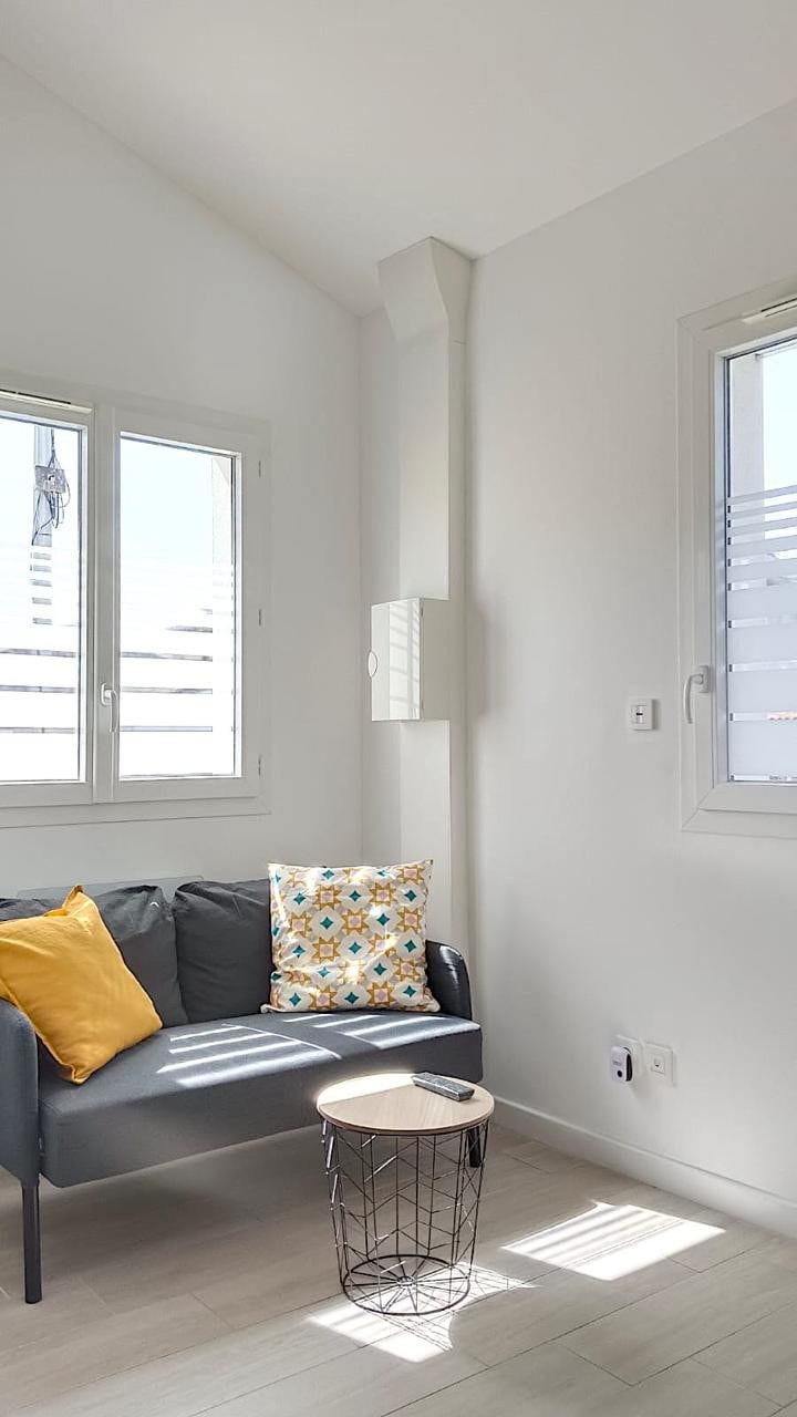 A cozy sitting area is highlighted by a modern gray couch adorned with a decorative pillow featuring a colorful pattern. A small round coffee table is positioned nearby. Natural light enters through two windows adorned with horizontal blinds.