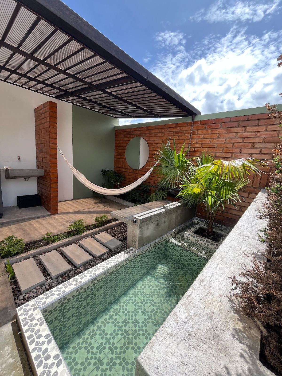 A private patio is showcased, featuring a small pond surrounded by pebbles and plantings. A hammock is suspended between structural supports, and a grill area is present on one side. The overhead covering provides shade while allowing natural light to filter through.