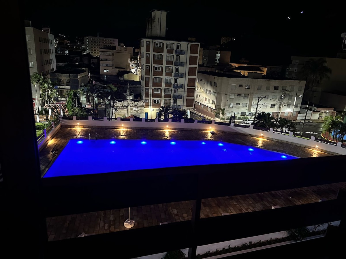 An evening view of a well-lit pool area, showcasing the illuminated water in a calming blue hue. Surrounding greenery and buildings are visible in the background, providing a sense of the vibrant city atmosphere.