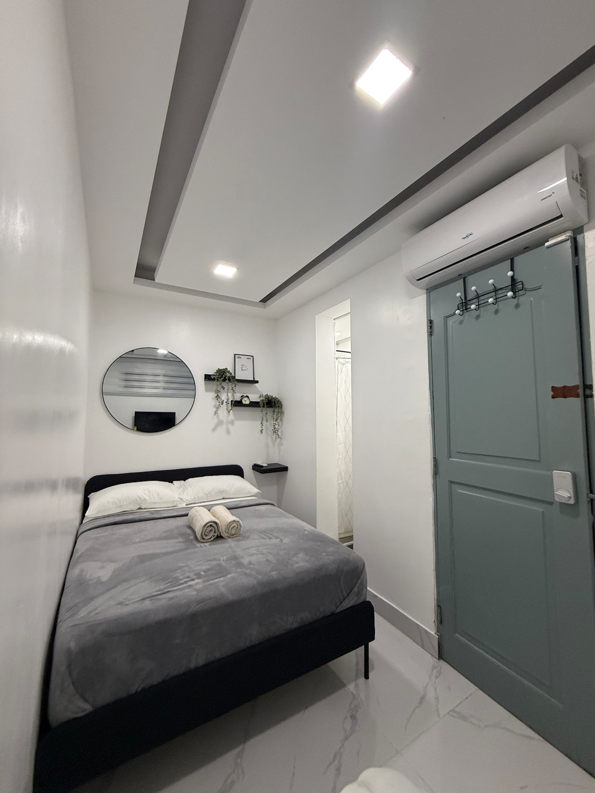 A compact bedroom features a comfortable bed with soft gray linens and freshly folded towels. A round mirror is mounted on the wall, while decorative plants add a touch of greenery. An air conditioning unit and a door with a sleek design are visible.