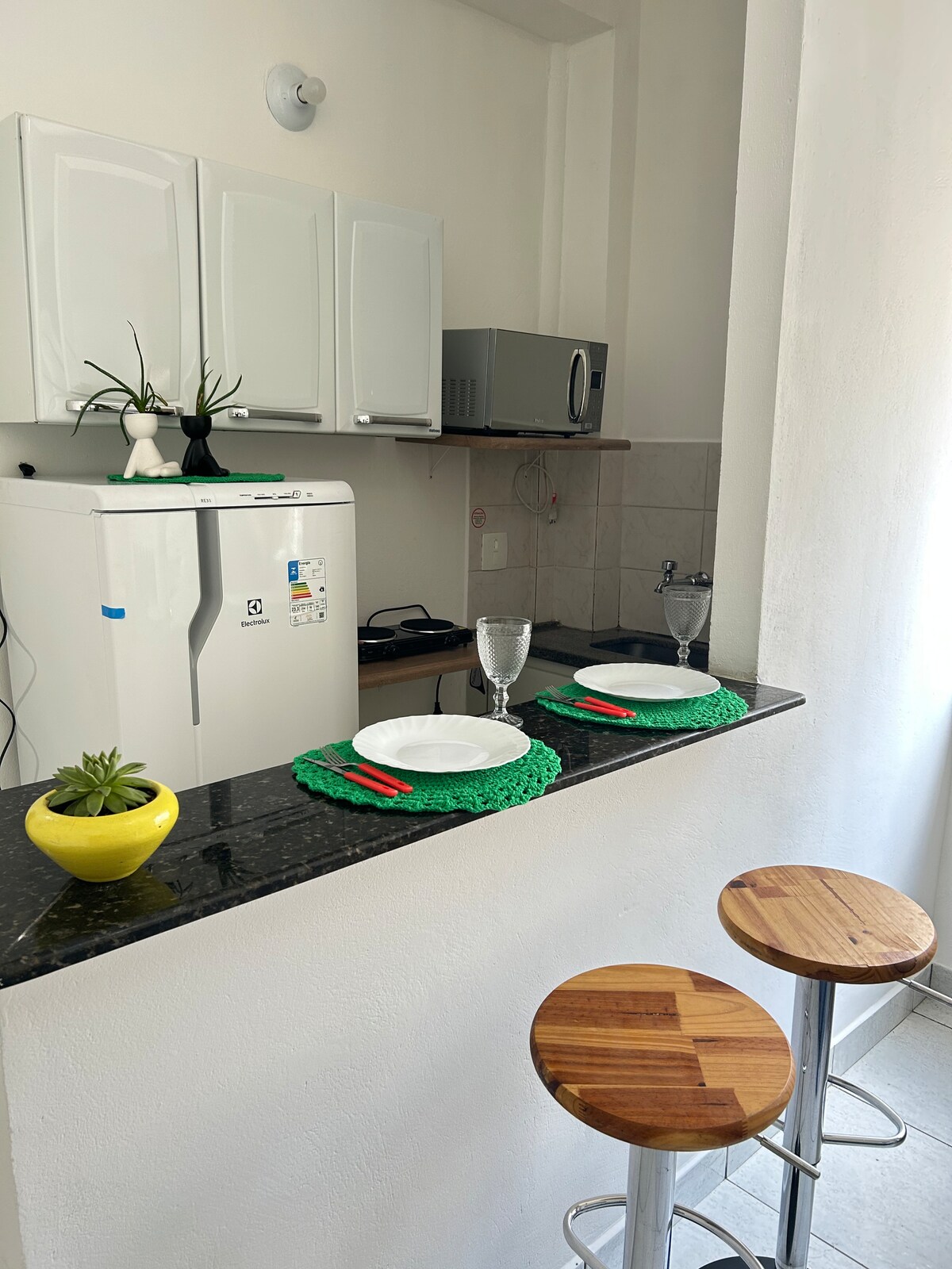 A compact kitchen area features modern white cabinetry and a stainless steel microwave. A black granite countertop is adorned with two place settings, including plates and glasses. Two wooden bar stools provide casual seating, complemented by a small green plant in a yellow pot.
