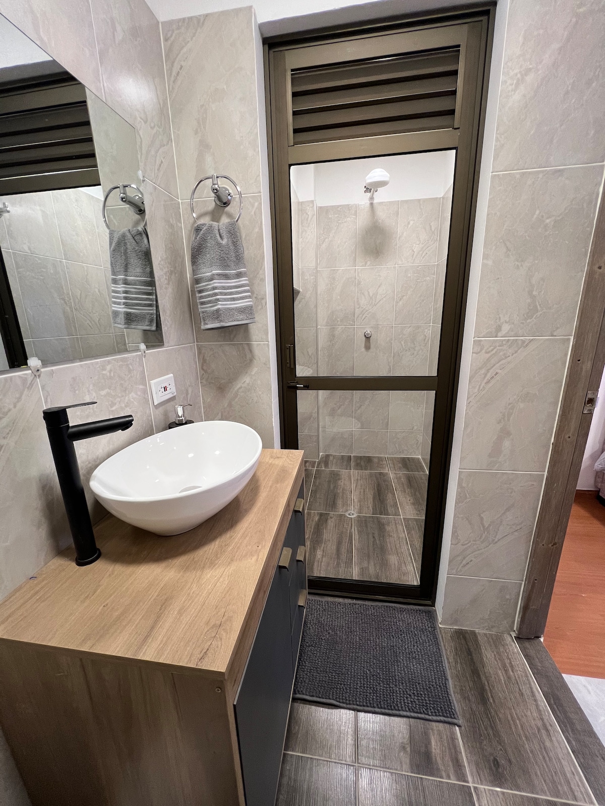 A modern bathroom features a sleek, oval sink atop a wooden vanity. A mirror reflects the surroundings, with two gray towels hanging nearby. A glass-walled shower stall is visible at the rear, showcasing tiled walls and a showerhead mounted above.