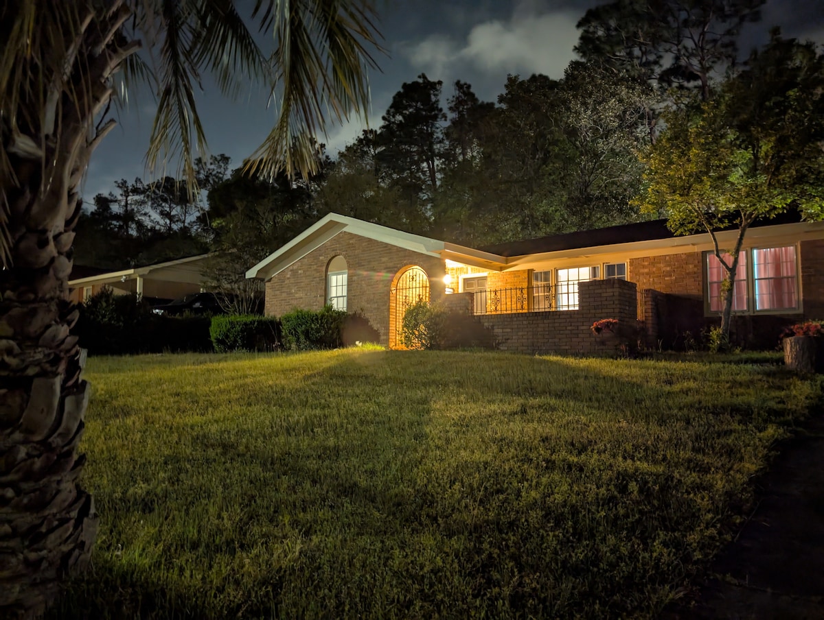 An inviting brick facade is illuminated by soft lighting, showcasing an archway entrance. Lush green grass surrounds the home, complemented by nearby palm trees and ambient tree foliage. Large windows allow glimpses of warm light from within, enhancing the serene nighttime atmosphere.