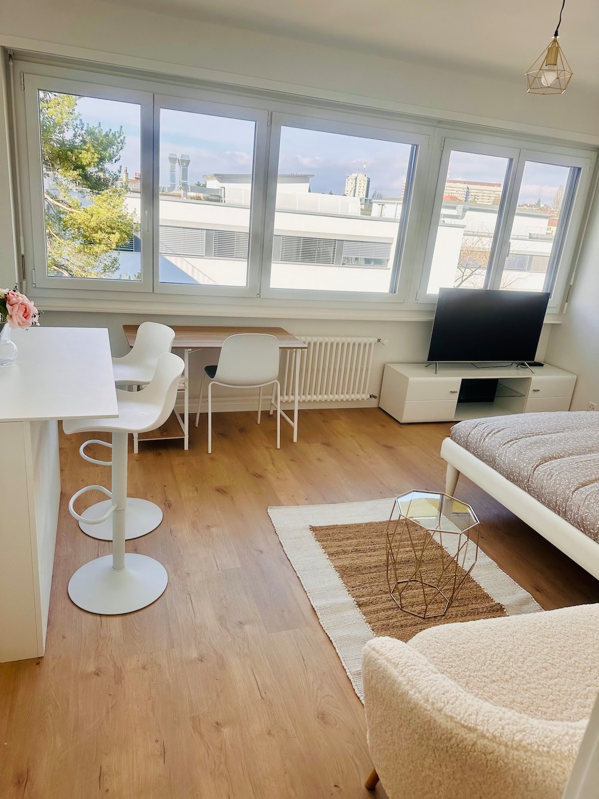 A bright living area is filled with natural light from large windows. A spacious bed is complemented by a white table and two chairs positioned nearby. A television is placed on a modern console unit, while a cozy rug adds warmth to the wooden flooring.