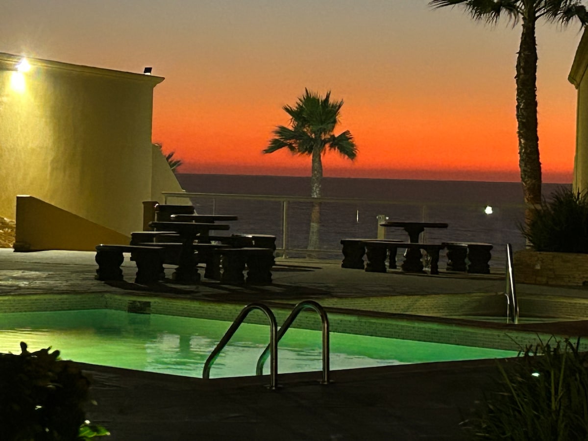 The image captures a serene pool area illuminated by soft lighting, with a view of a vibrant sunset casting hues of orange and purple in the sky. Palm trees are visible in the foreground, enhancing the tranquil seaside setting.
