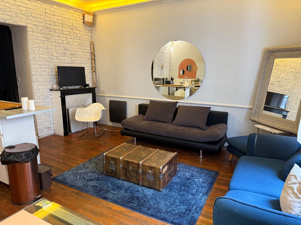 The living area features a combination of seating, including a dark sofa bed and a standard blue couch. A rustic coffee table sits in front, while a large mirror reflects the space. A TV and a minimalist white chair are also visible.