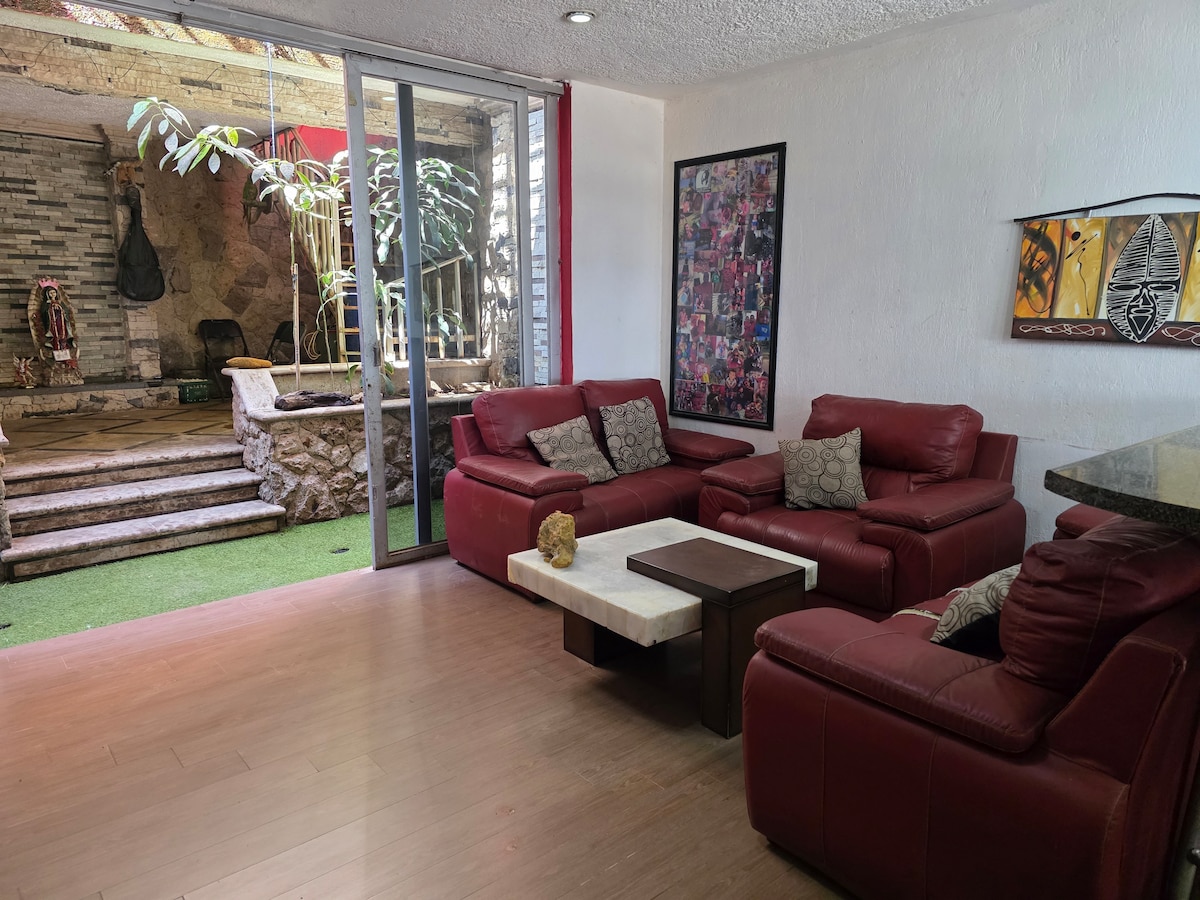 A living area features a set of red leather sofas arranged around a light-colored coffee table. Large glass doors provide a view of an outdoor area with greenery and stone steps, creating a seamless connection to nature.