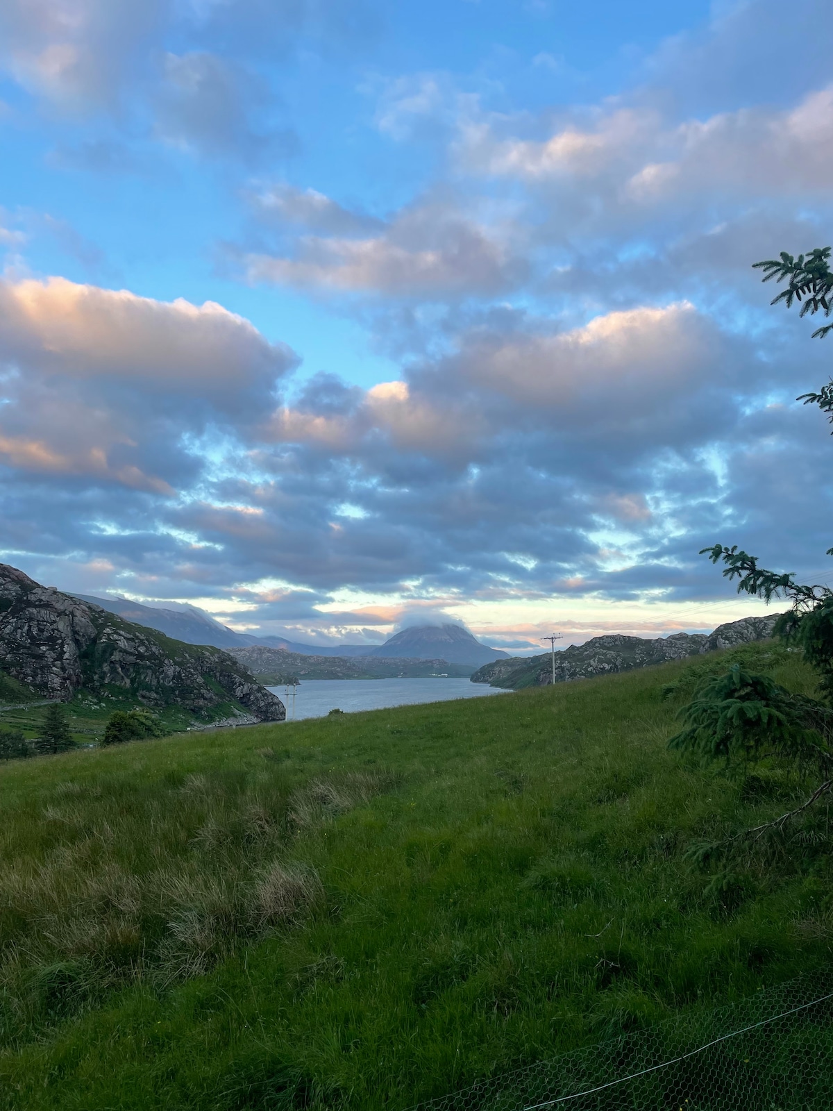 A scenic view showcases rolling green hills leading to a serene body of water, framed by dramatic mountains. The sky is filled with soft clouds bathed in warm hues, creating a peaceful atmosphere in the natural landscape.