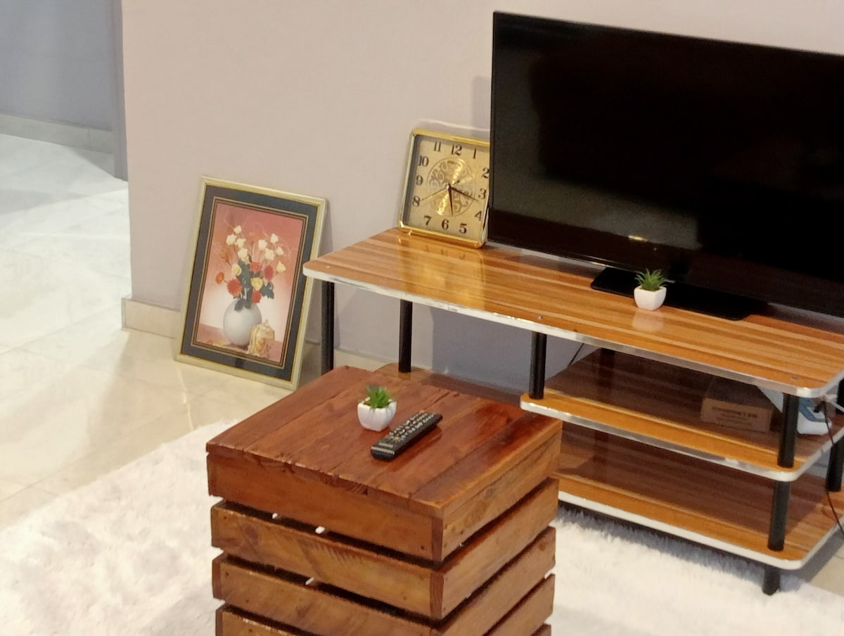 A wooden coffee table with a rustic design sits in front of a sleek TV stand. A small potted plant decorates the table, complemented by a remote control. A vintage clock and framed artwork featuring floral arrangements are positioned nearby.