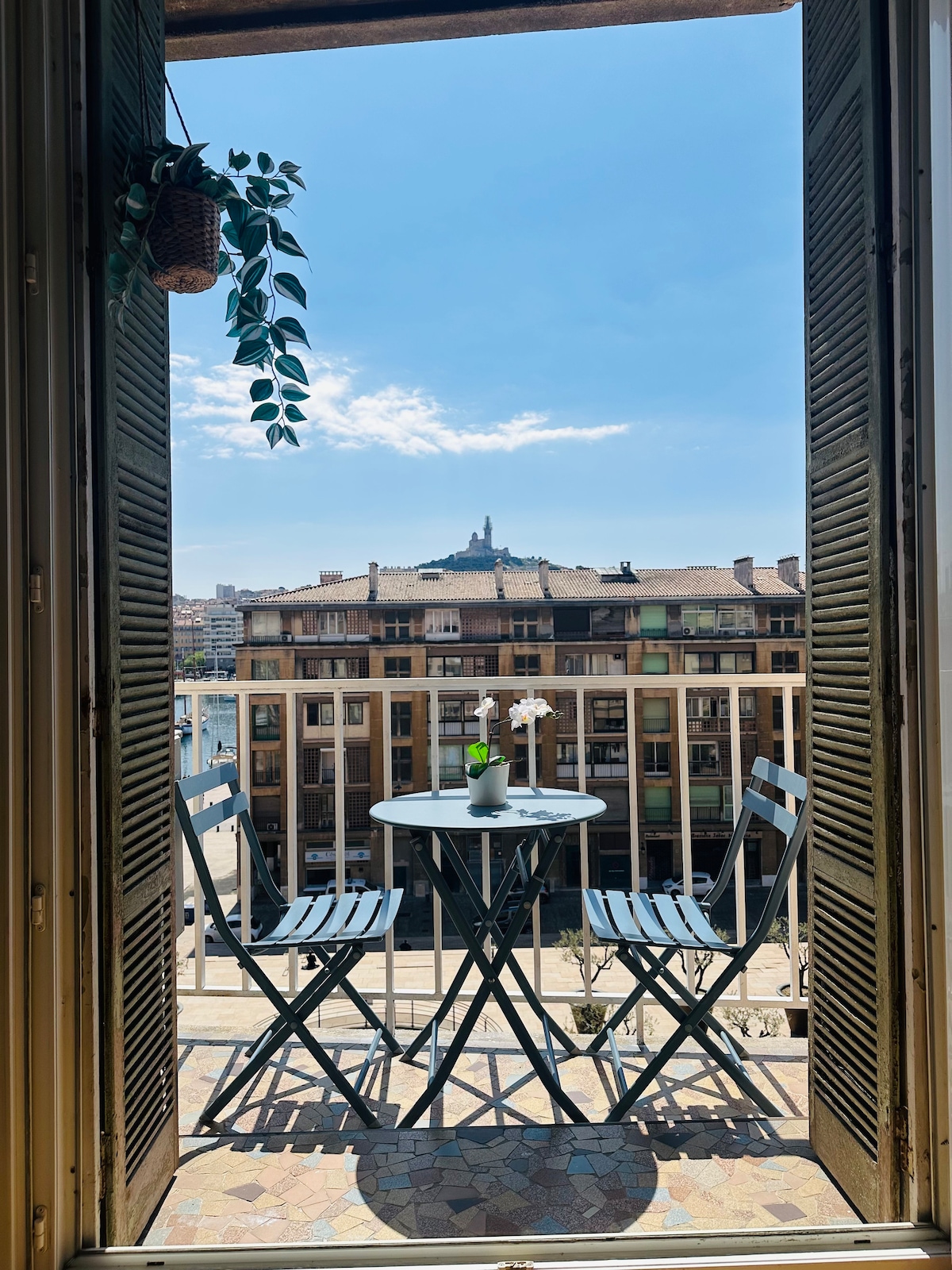 A small balcony is visible, featuring a round table and two folding chairs, with a potted plant hanging nearby. The view overlooks a bustling urban landscape, with Notre-Dame-de-la-Garde prominently positioned in the background against a bright blue sky.