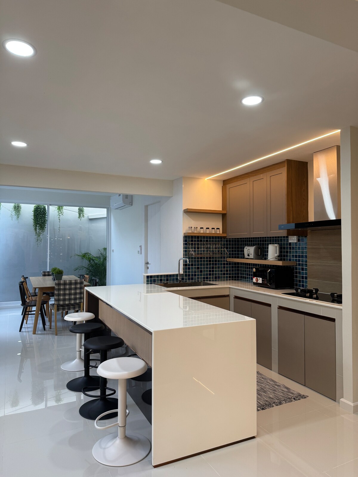 A modern kitchen features a long, sleek countertop with seating options on one side. Contemporary cabinets complement tile backsplash, while appliances are neatly arranged. A dining area with a wooden table and chairs is visible through a glass door, adding a touch of greenery to the space.