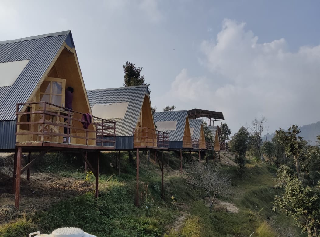 A row of A-frame wooden cottages is set on stilts, positioned against a backdrop of trees. Each cottage features a spacious deck with wooden railings, offering unobstructed views of the surrounding landscape and distant hills under a clear sky.